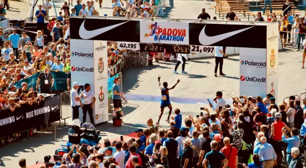 Meta-brama z napisem "Padova Marathon" i biegnącym zawodnikiem, tłum widzów, zegar z czasem.